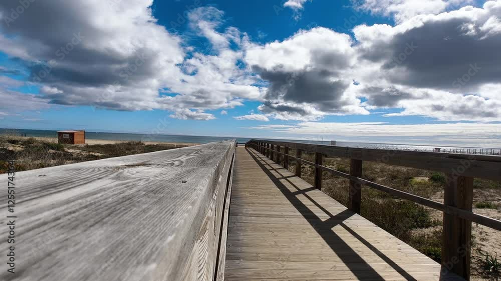 A wooden boardwalk stretches out towards leading to the beach, a blue sky filled with fluffy white clouds. The beach and ocean can be seen in the distance. In Algarve, Portugal.