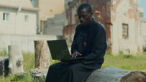 Modern African American priest dressed in black cassock working on laptop while sitting on log on summer day photo