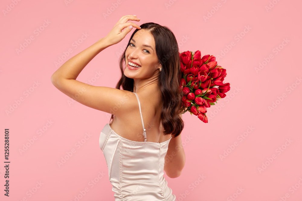 Glad pretty woman holding bouquet of red flowers and smiling at camera, posing in elegant dress on pink background. Women's day celebration Glad pretty woman holding bouquet of red flowers and smiling at camera, posing in elegant dress on pink background. Women's day celebration