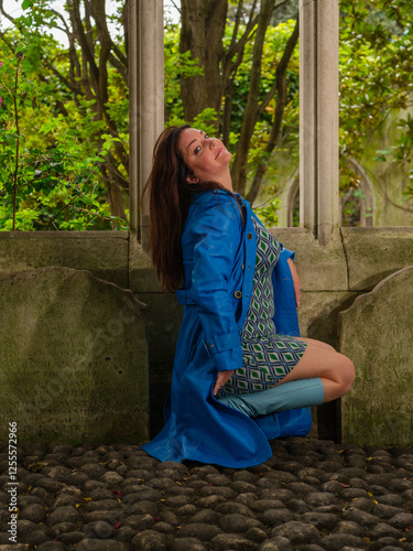 A woman in a blue coat, patterned dress, and matching boots poses playfully on cobblestone ground in front of an old stone window frame with greenery