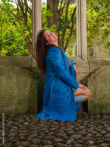 A woman in a blue coat, patterned dress, and matching boots poses playfully on cobblestone ground in front of an old stone window frame with greenery