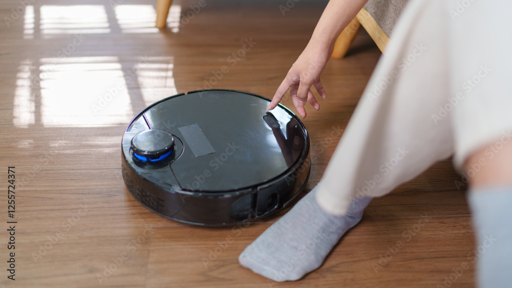 Asian woman turning on a robotic vacuum while sitting on a sofa in a cozy living room Asian woman turning on a robotic vacuum while sitting on a sofa in a cozy living room