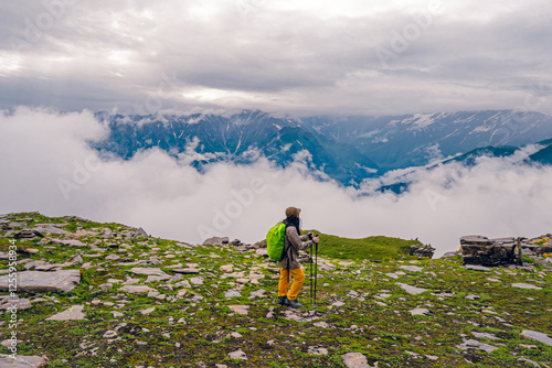 On a cloudy day, a woman in hiking clothes stands on green grass hill in Bhrigu lake trek, Manali, India. There are mountains with snow on top. Idia for adventure activity background with copy space.