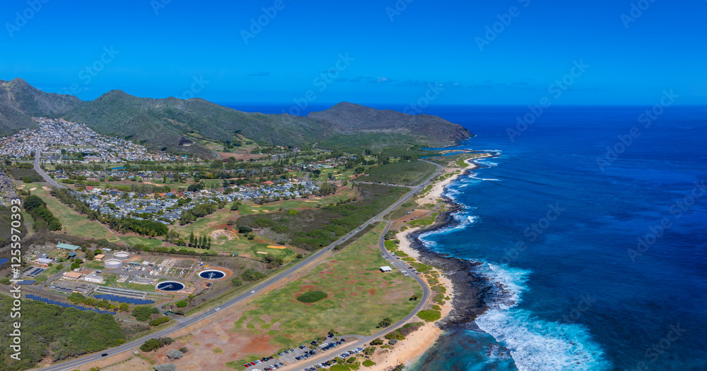 Aerial view of Oahu, Hawaii, showing a highway along the coast, lush greenery, a small beach, water treatment structures, and a residential area by mountains. Aerial view of Oahu, Hawaii, showing a highway along the coast, lush greenery, a small beach, water treatment structures, and a residential area by mountains.