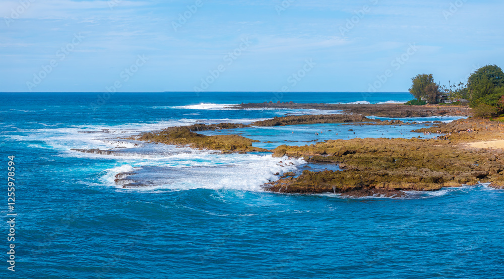 Rugged rocky coastline of Oahu, Hawaii, with blue ocean waves, golden sandy patches, and lush green vegetation under bright, sunny weather.