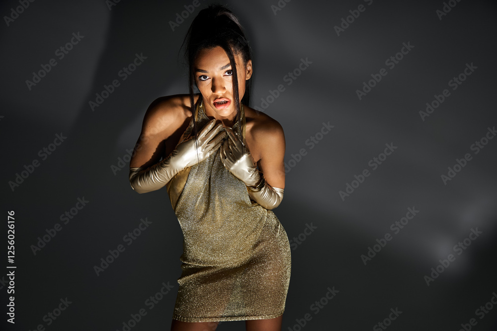 Obraz premium Elegant young woman showcases trendy fashion in a glamorous golden attire under soft lighting