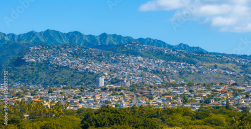 Photography Aerial view of a hillside residential area on Oahu island, Hawaii, with lush green mountains, dense housing, palm trees, and a clear blue sky
