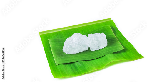 Pieces of white alum sit on a banana leaf, set against a white background. This simple yet meaningful display represents the use of natural minerals in purification, skincare, and household remedies.