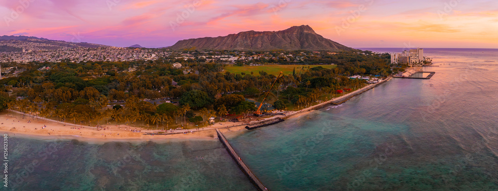 Obraz premium Aerial view of Waikiki Beach in Honolulu, Hawaii, with Diamond Head Crater in the background, sandy shores, turquoise waters, and urban structures at sunset.