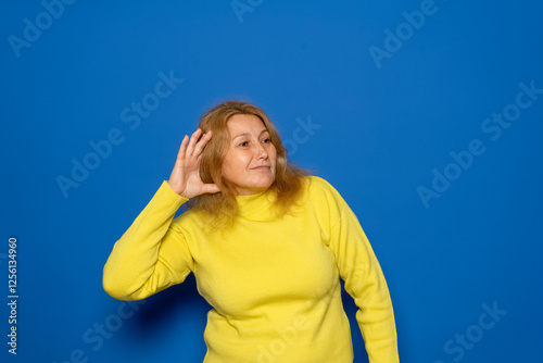 Caucasian woman in her 40s wearing a yellow sweater with one hand on her ear eavesdropping on something, isolated over blue background. Gossip and rumors concept