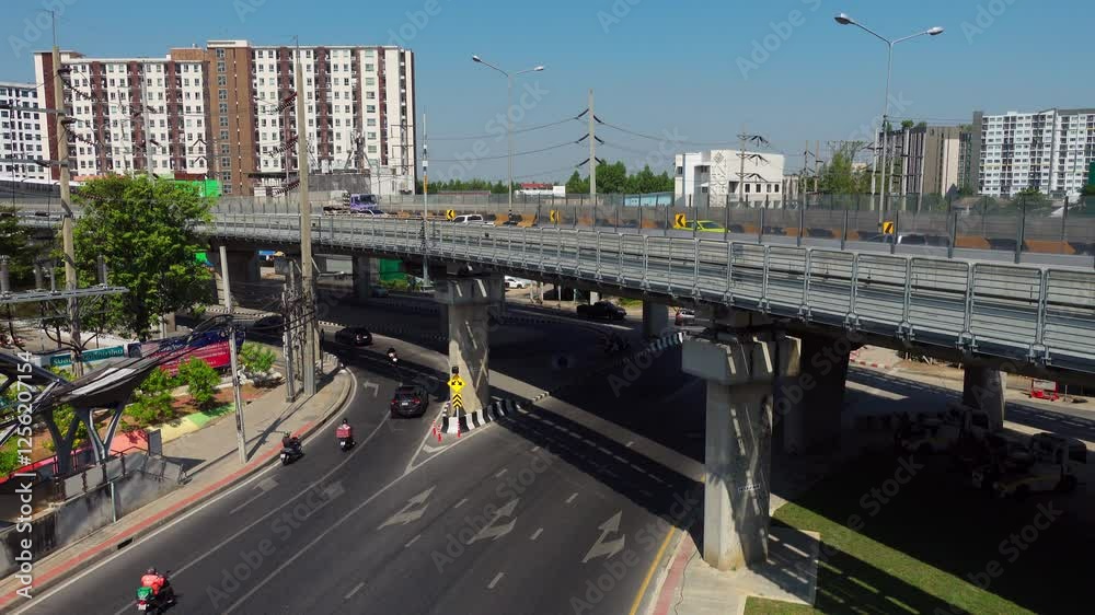 Aerial view showcasing modern urban transportation in a bustling city. An elevated train moves along its track while cars and motorcycles navigate the circular intersection below. The scene captures t