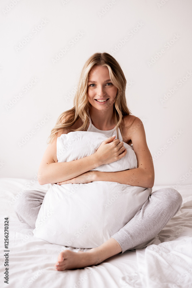 young blonde woman hugging white pillow sitting on cozy bed looking in bedroom. young blonde woman hugging white pillow sitting on cozy bed looking in bedroom.