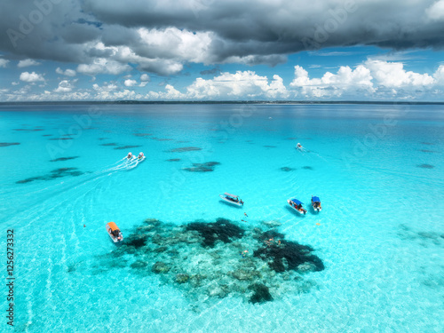 Aerial view of colorful boats and yachts in clear water on sunny summer day. Mnemba island, Zanzibar. Top view of sandbank in low tide, blue sea, swimming people, yachts, dramatic sky with clouds