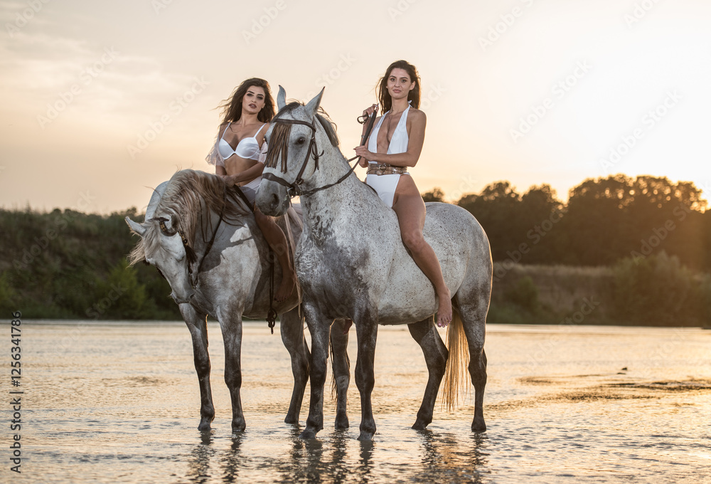 Beautiful brunettes woman with long hair ,white bikini and white shirt rides on a white horse in the river. Sensual and attractive girls interacting and rides a horse in the water at sunset