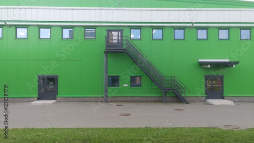 An industrial building with square windows, with several entrances at the bottom and one entrance on the second floor, reached by a metal staircase with handrails, is located near a grassy lawn photo