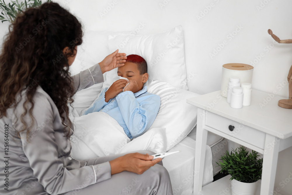 Fototapeta premium Sick little African-American boy and his mother with thermometer lying in bedroom