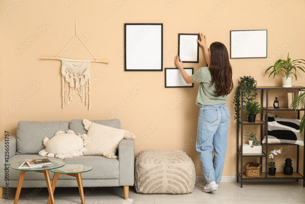 Young woman hanging blank frame on beige wall in living room. Back view Young woman hanging blank frame on beige wall in living room. Back view