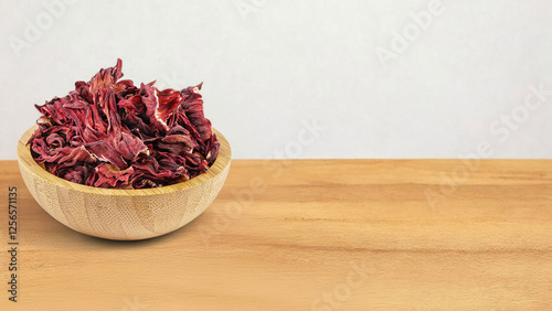 A wooden bowl filled with vibrant red dried Hibiscus sabdariffa leaves rests on a wooden surface. The deep red leaves crinkle, displaying their rich color and textured appearance.
 photo