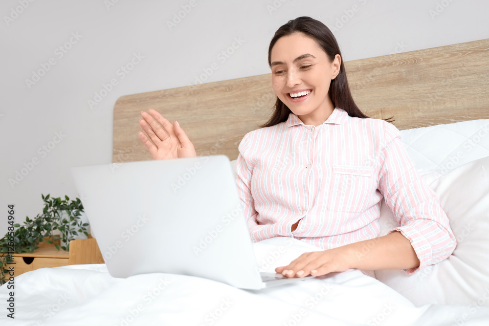 Young woman in striped pajamas with laptop video chatting on bed at home Young woman in striped pajamas with laptop video chatting on bed at home