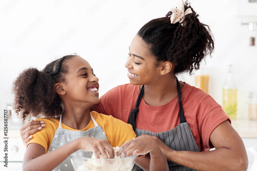 Cute smiling african american young mother and daughter teenager cuddling at kitchen while baking, black mom hugging her happy kid making dough for pastry, wearing aprons, copy space Cute smiling african american young mother and daughter teenager cuddling at kitchen while baking, black mom hugging her happy kid making dough for pastry, wearing aprons, copy space