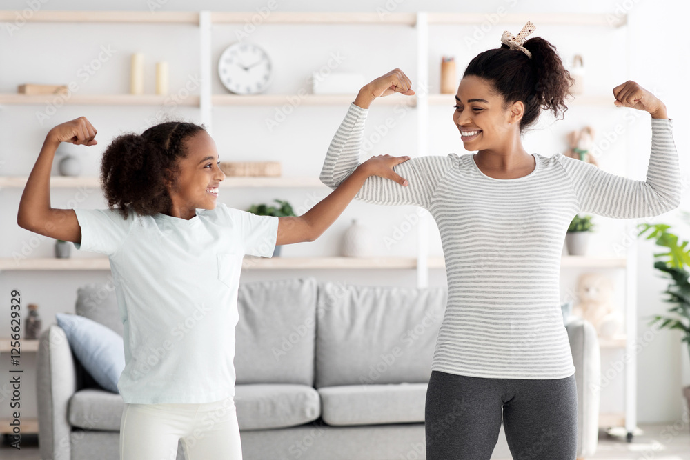 Strong african american mother and teen daughter in sportswear showing their muscles while doing morning exercises together at home, athletic black family demonstrating biceps and smiling Strong african american mother and teen daughter in sportswear showing their muscles while doing morning exercises together at home, athletic black family demonstrating biceps and smiling