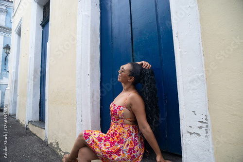 Beautiful young Caucasian woman with braids and colorful dress sitting in front of a blue door of an old house posing for a photo. Confident person. photo