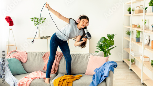 Cheerful Indian teen girl with blowdryer as mic singing her favorite song and dancing on sofa at home, full length. Cool teenager pretending to be famous singer, having fun in messy room