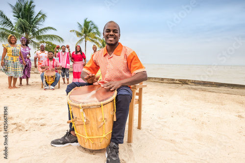 Young Latino man of the Garifuna ethnic group plays the drum with joy, his friends dance in the background.