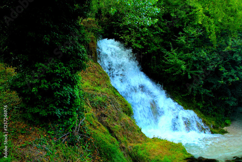 Energizing view from Sefro (Macerata) with a lush waterfall streaming down the slope and collecting into a rivulet below, its creamy white waters surrounded by thriving greenery and fresh dark foliage photo