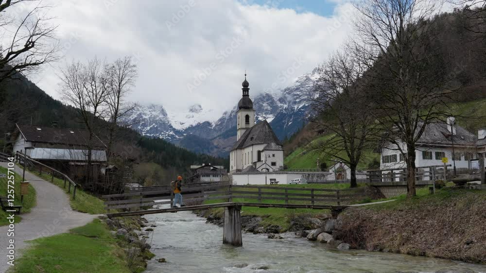 A local man walking through traditional wooden bridge over river stream with parish church of St Sebastian in back in Bavarian alps foothills, Europe A local man walking through traditional wooden bridge over river stream with parish church of St Sebastian in back in Bavarian alps foothills, Europe