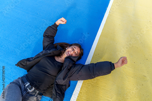 Happy young woman lying on blue and yellow ground raising fists celebrating success, expressing joy and excitement photo