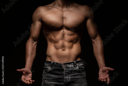 Muscular man's torso with defined abs, hands on hips, wearing jeans and a bracelet in a studio with dark background
