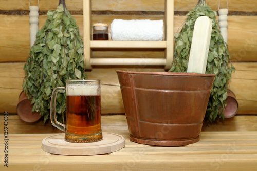 A glass mug of light beer stands on a plank bench in the sauna interior next to a copper vat for sauna treatments against a log wall with dry birch brooms, copper ladles and a shelf with a bath towel.