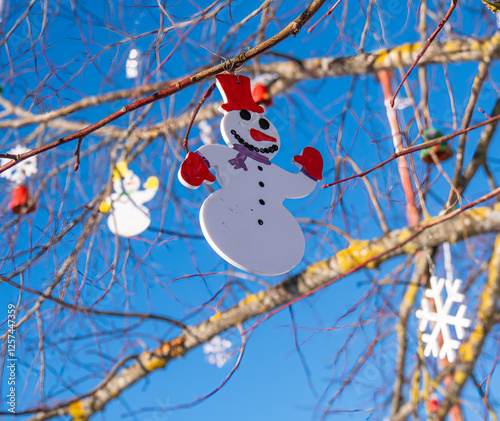 A cheerful snowman ornament hangs on a tree branch against a clear blue sky photo