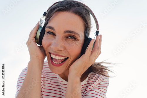 Cheerful woman listening music through wireless headphones under clear sky