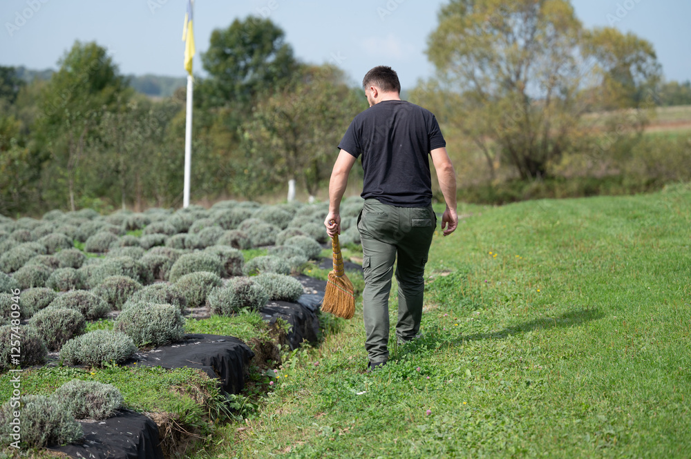 Fototapeta premium A gardener walks away from rows of garden plots while carrying a broom. The scene is set on a sunny day with trees, grass and a flag in the background. Landscaping and groundskeeping.