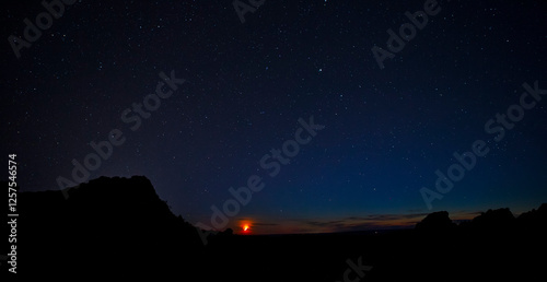 The Gobi Desert night sky is filled with stars as the fiery red sun sets over the horizon, creating a stunning contrast between the vast, dark sky and the glowing desert landscape.
