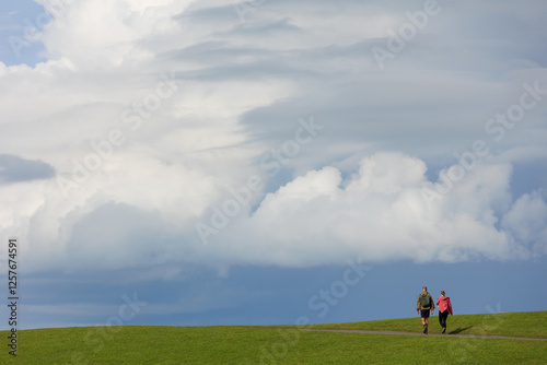 View of two people walking, seen from the back in front of a lot of cloudy sky and and a bit of grass like they are walking straight into heaven