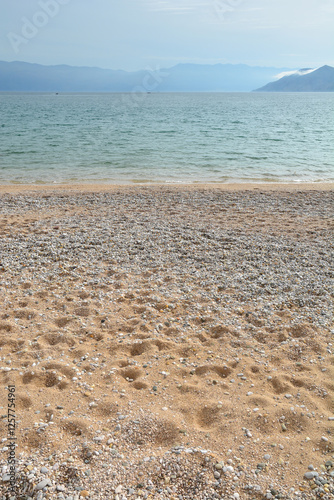 Fototapeta Naklejka Na Ścianę i Meble -  A serene late autumn scene of Baska beach featuring a mix of pebble and sandy shoreline, with the Velebit mountain and Prvic island visible in the distance under a tranquil sky