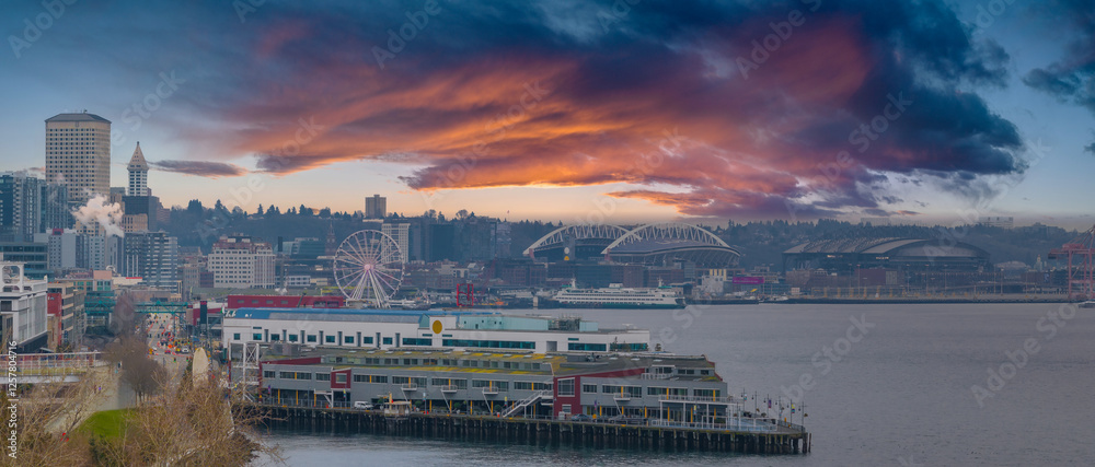 Obraz premium Aerial view of Seattle, Washington, featuring the Great Wheel, Smith Tower, Lumen Field, T Mobile Park, a pier, a cargo ship, and a vibrant sky.