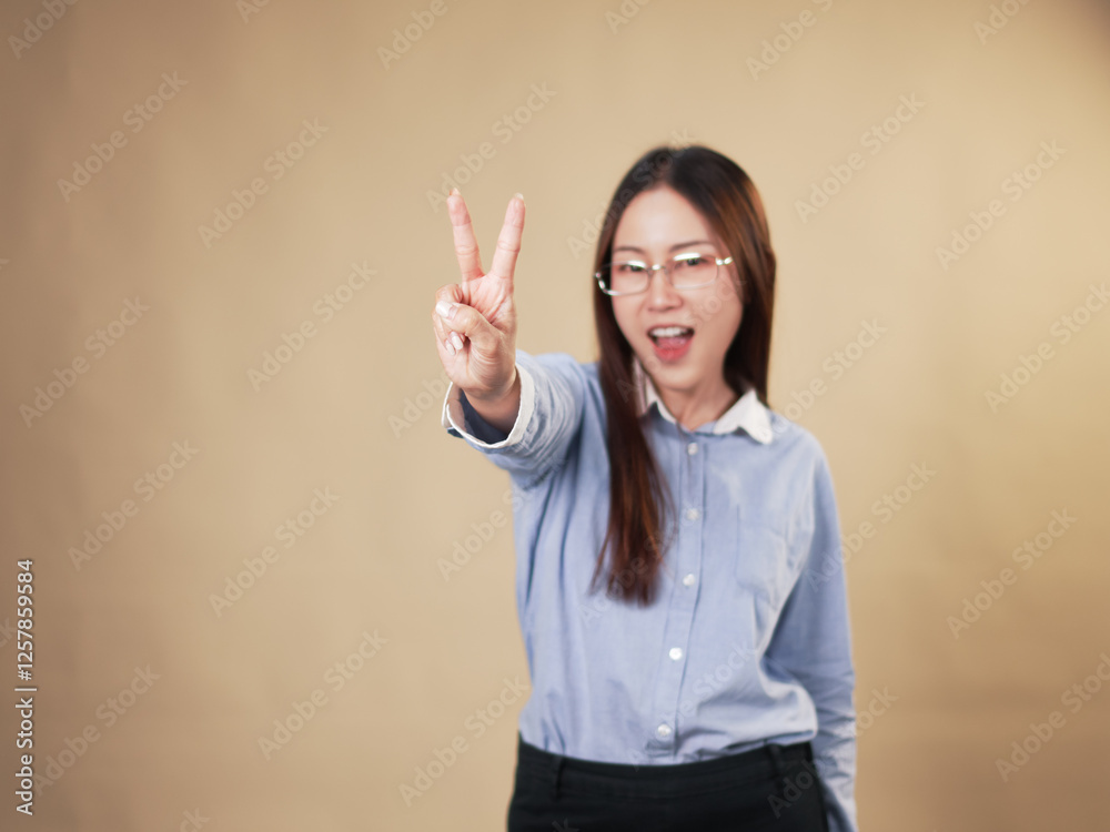 A young woman in glasses and a light blue shirt is smiling and making a peace sign or victory sign with her hand, expressing joy and confidence A young woman in glasses and a light blue shirt is smiling and making a peace sign or victory sign with her hand, expressing joy and confidence