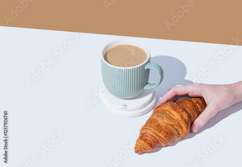 Hand holding fresh croissant on white broun background and cup of coffee with milk.