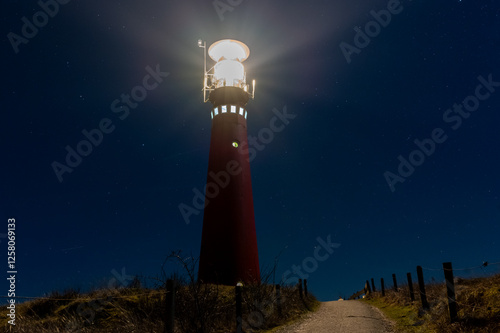The red lighthouse on Schiermonnikoog is lit up at night. The light is shining brightly. The lighthouse is on a hill