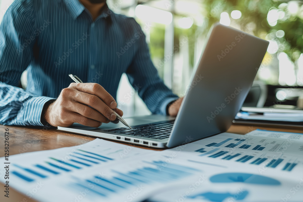 Man working on laptop with papers scattered on a table in a modern office setting Man working on laptop with papers scattered on a table in a modern office setting