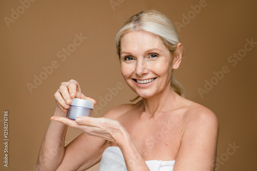 Glad lively European woman of middle age applies anti aging cream on face has natural beauty cares about skin in white towel holds jar of cosmetic product looks happily at camera poses indoor