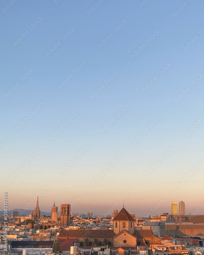Fototapeta premium Panoramic view of Barcelona at sunset. The warm-colored skyline is dominated by a mixture of historic and modern architecture, including church spires and skyscrapers, and contrasts with the blue sky