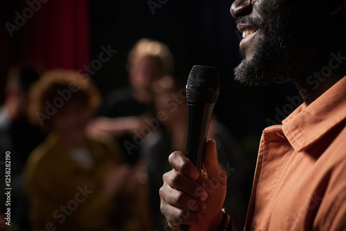 Man smiling while holding microphone during speech at an event with blurred audience in background focusing on microphone in hand