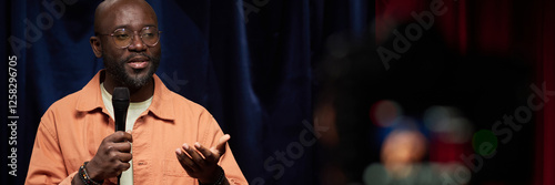 Konstfotografi African American man wearing glasses holding microphone while delivering speech on stage bathed in soft light