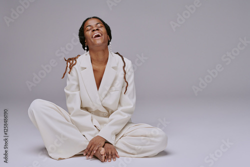 Young woman laughing while sitting cross-legged in a relaxed pose, wearing white attire against a neutral background photo