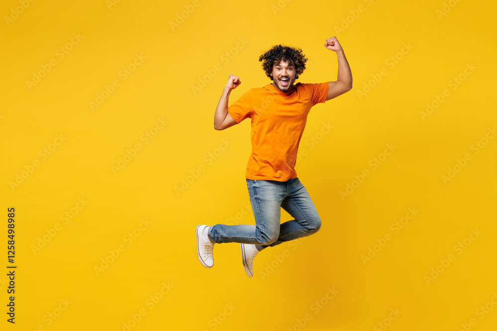Obraz premium Full body young happy fun overjoyed cheerful cool Indian man he wear orange t-shirt casual clothes doing winner gesture celebrate clenching fists say yes jump high isolated on plain yellow background.